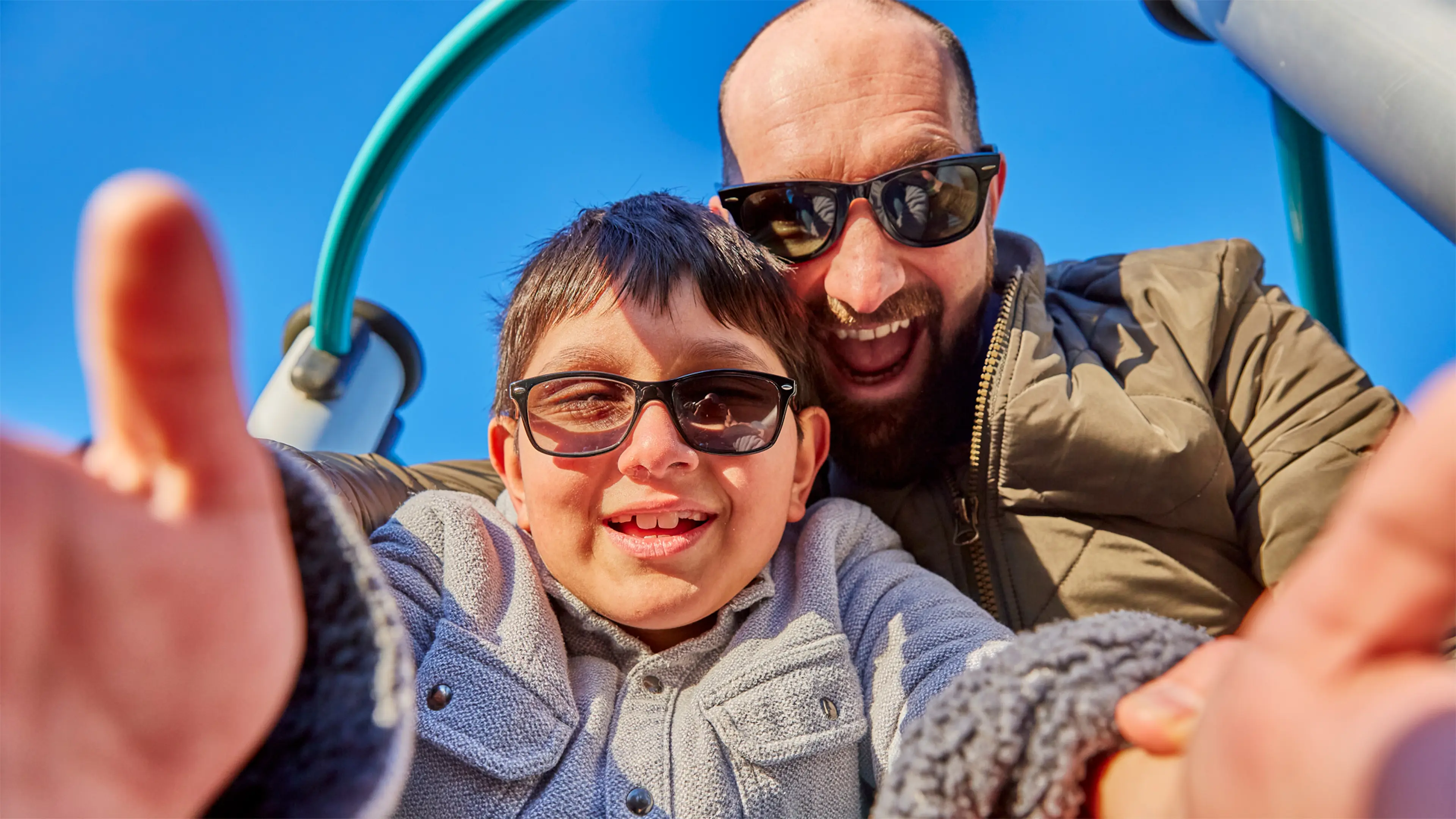Teddy, a child who has a vision impairment, takes a smiling selfie with his dad Phil, as they play in a playground.