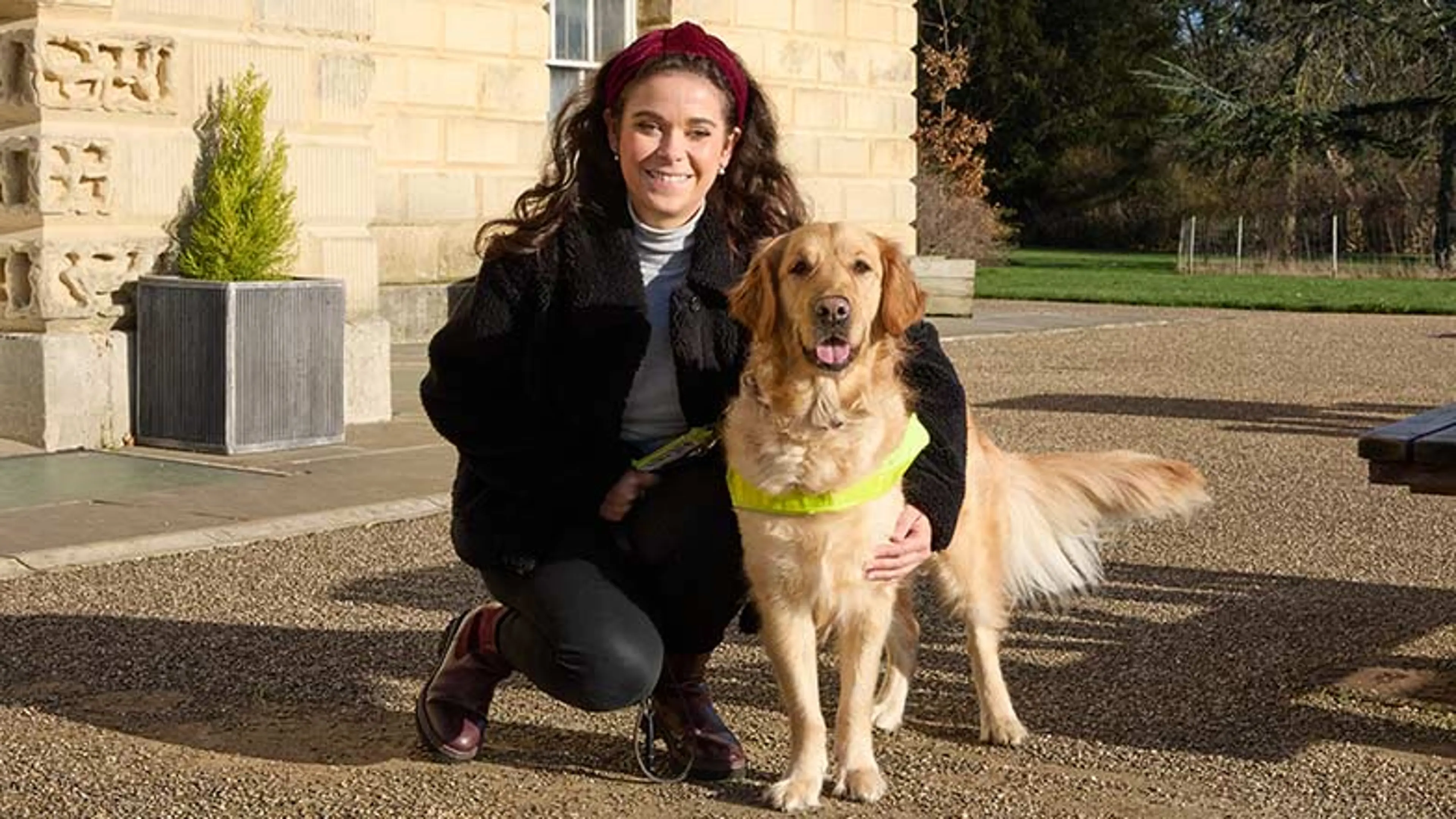 Guide dog owner Taylor crouching down next to guide dog Jilly in harness in the sunshine