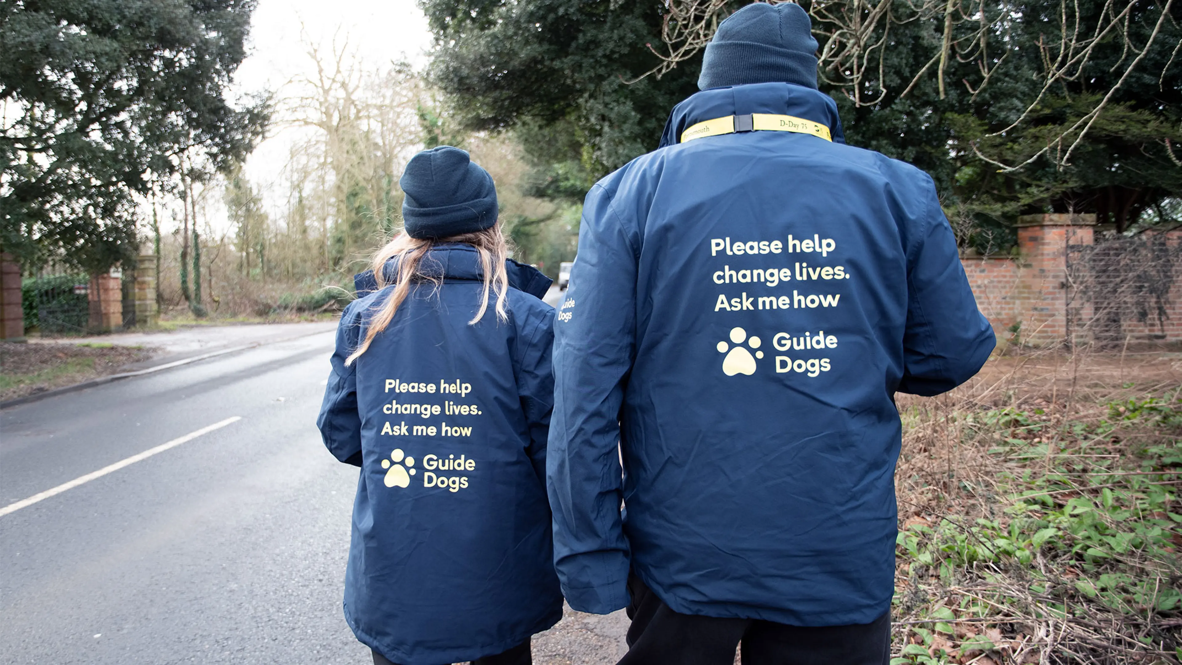 2 face to face fundraisers walking along a road facing away from the camera. Their jackets have 'Please help change lives. Ask me how' text on the back.