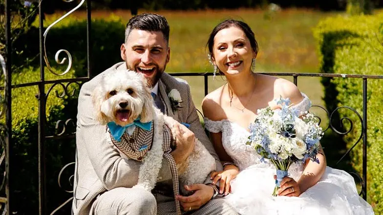 A bride and bridegroom sitting together smiling with a small dog on the bridegroom's lap. The dog is wearing a bow tie and waistcoat harness.
