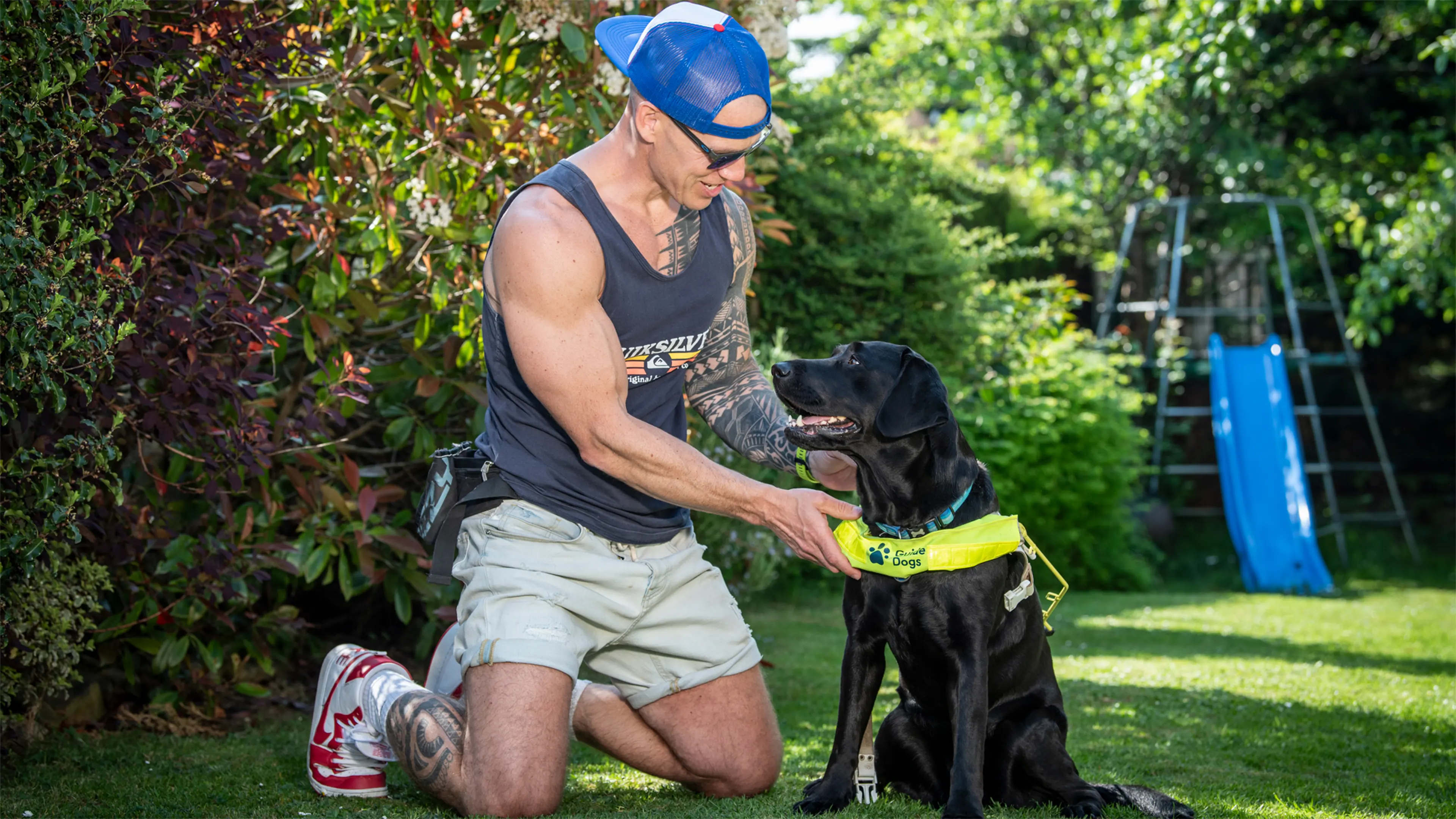 Personal trainer and guide dog owner Mark kneels in his garden next to Mary, his black Labrador guide dog.