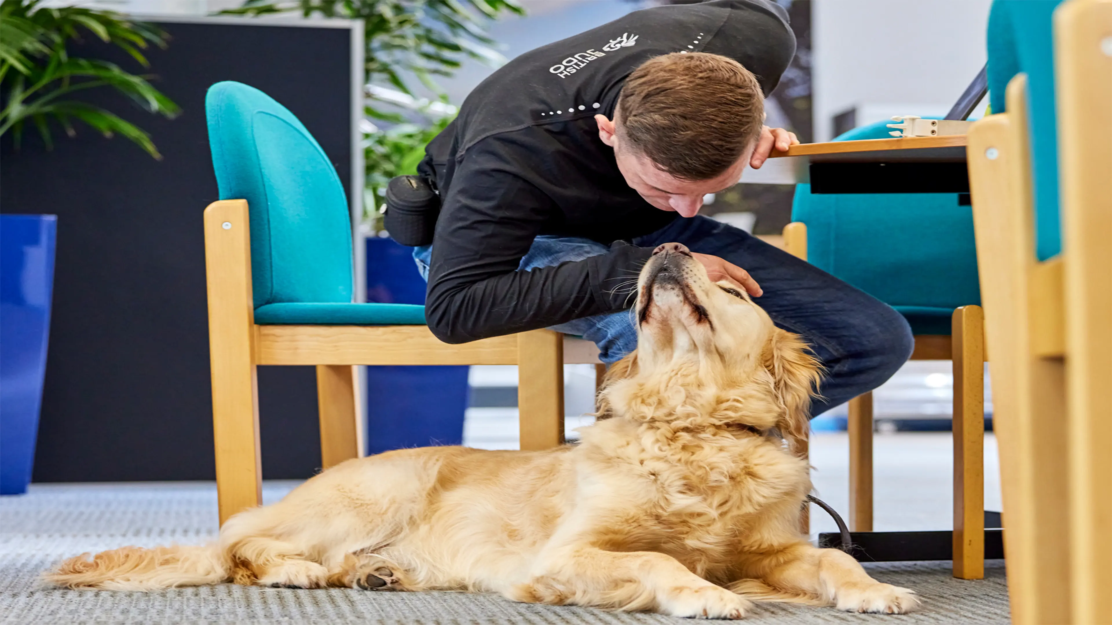 Guide dog owner Scott leans down from his chair to give guide dog Milo a scratch on the head, as he lies at his feet. 