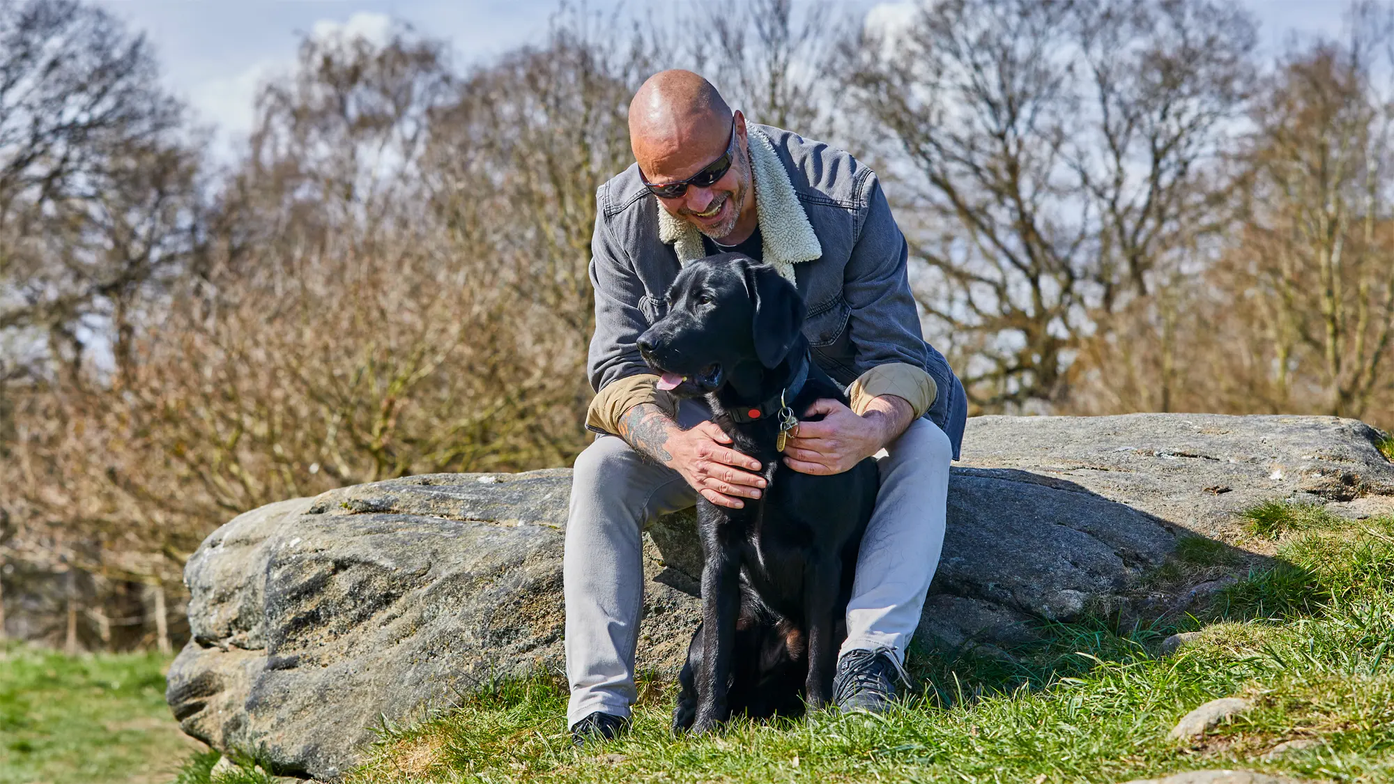 Guide dog owner Terry sits on a rock in a grassy park with his arms around guide dog Spencer.