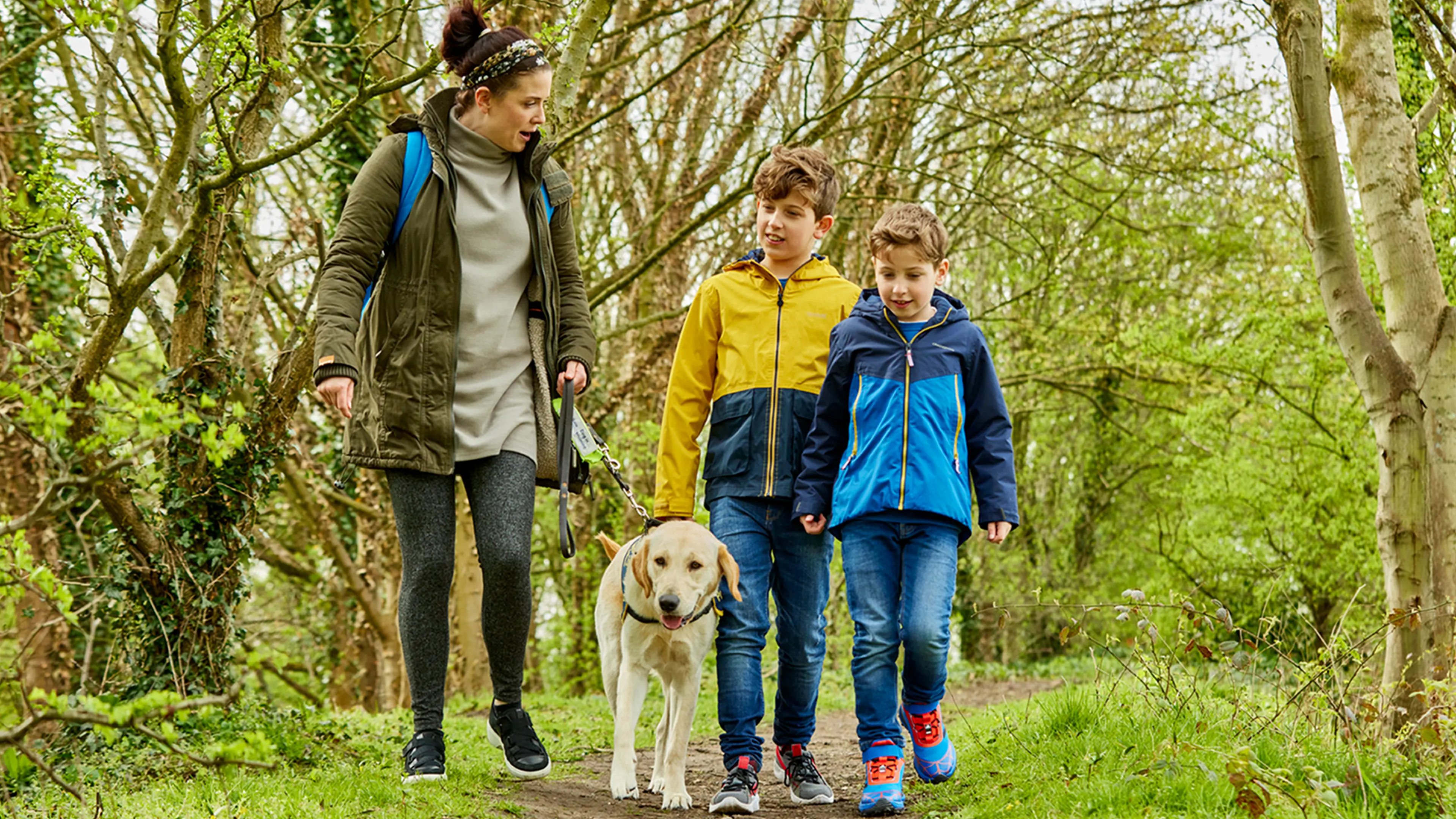 A Guide Dogs Puppy Raiser walks with her guide dog puppy and two children along a woodland path.