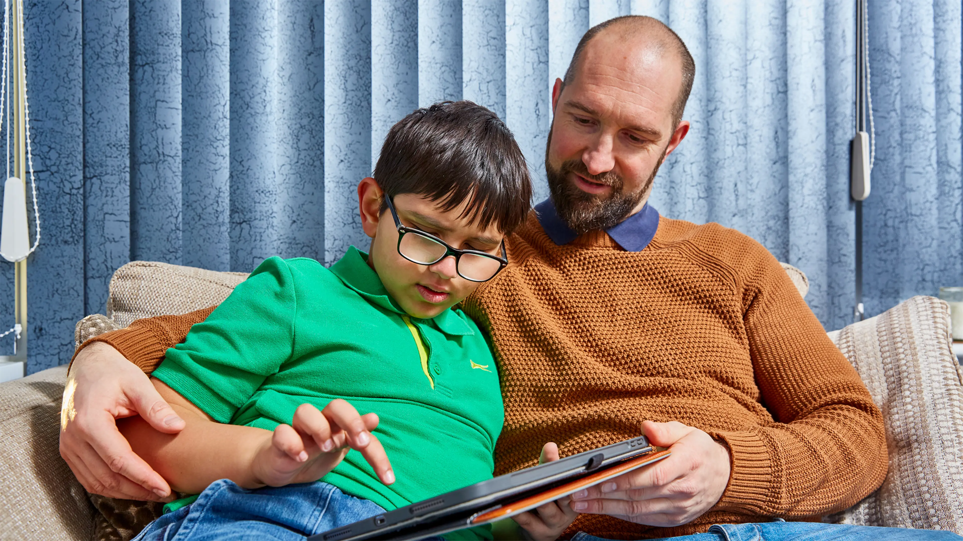 Teddy, a child who has a vision impairment, sits on a sofa with his dad. They are holding and interacting with a tablet.
