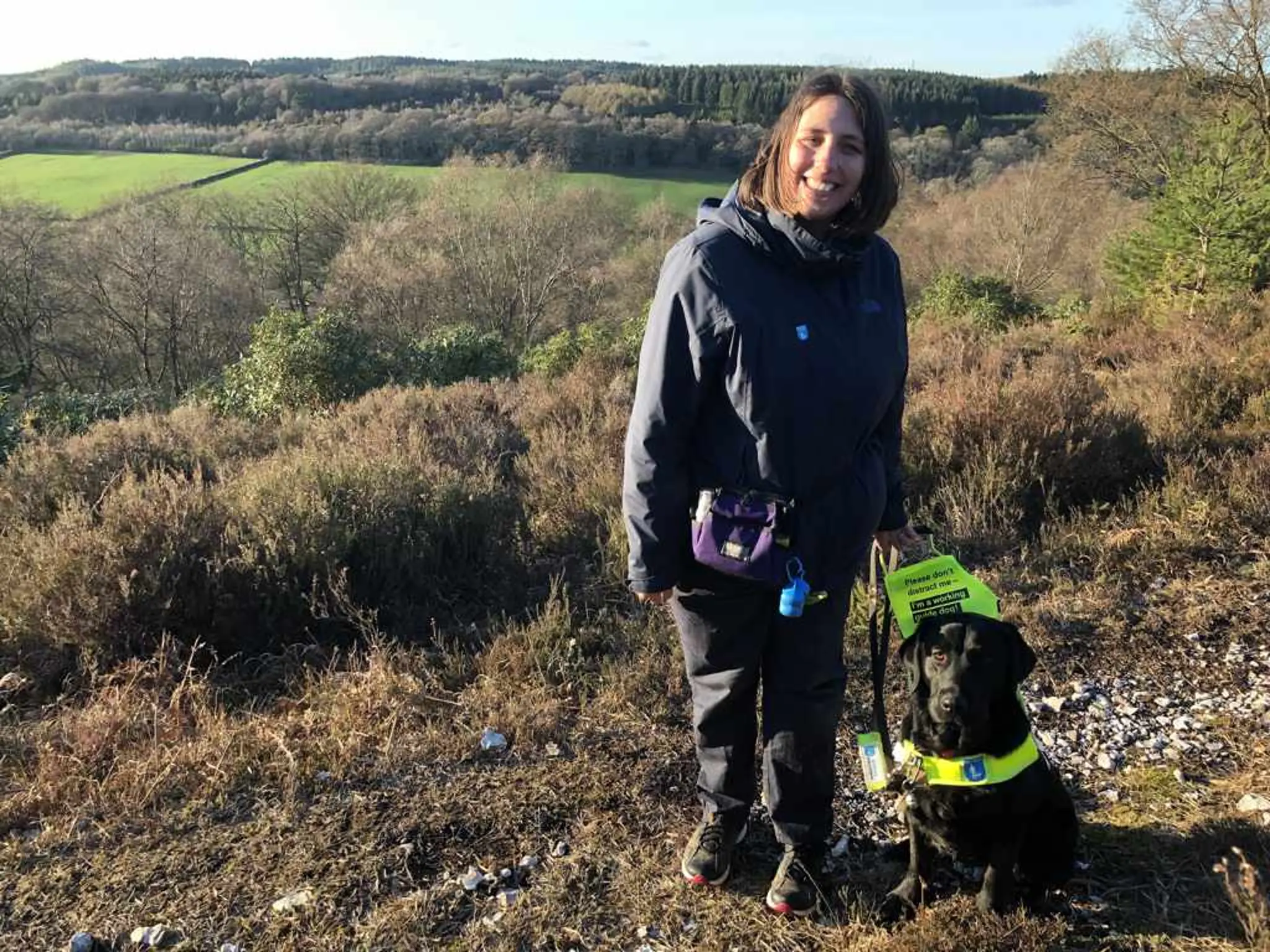 Guide dog owner Lauren stands and smiles in a field with guide dog Grace sat beside her in a harness