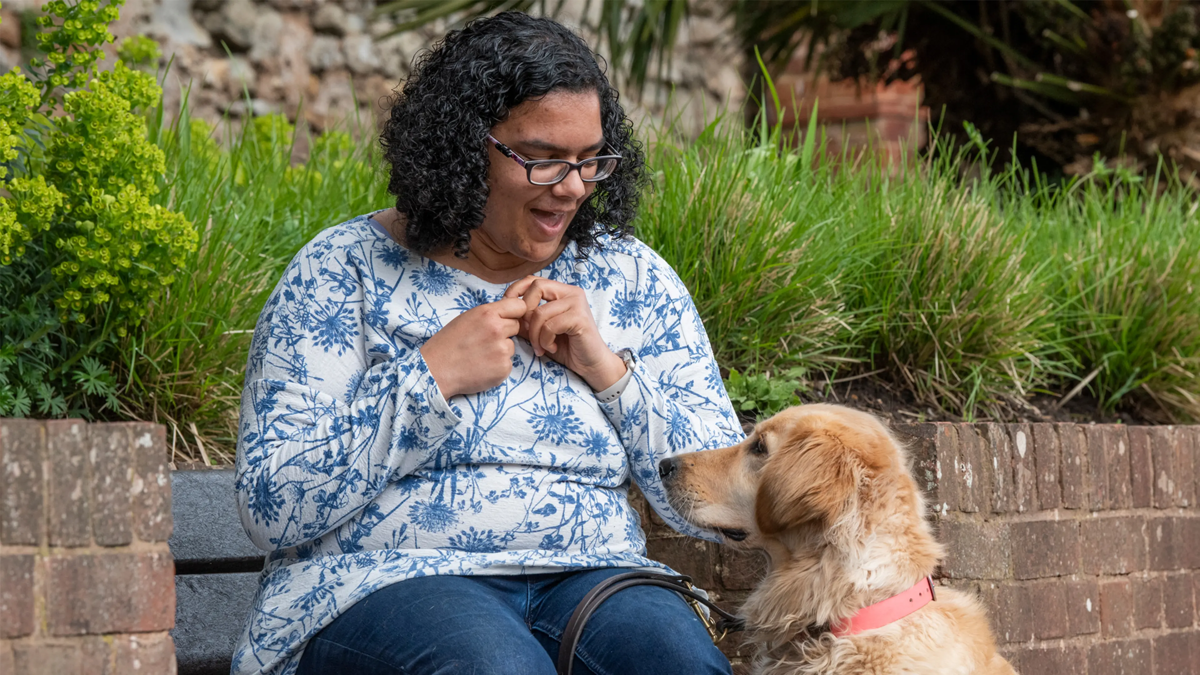 Guide dog owner Lena sits on a bench in a garden, looking down at her guide dog Alex who sits beside her.