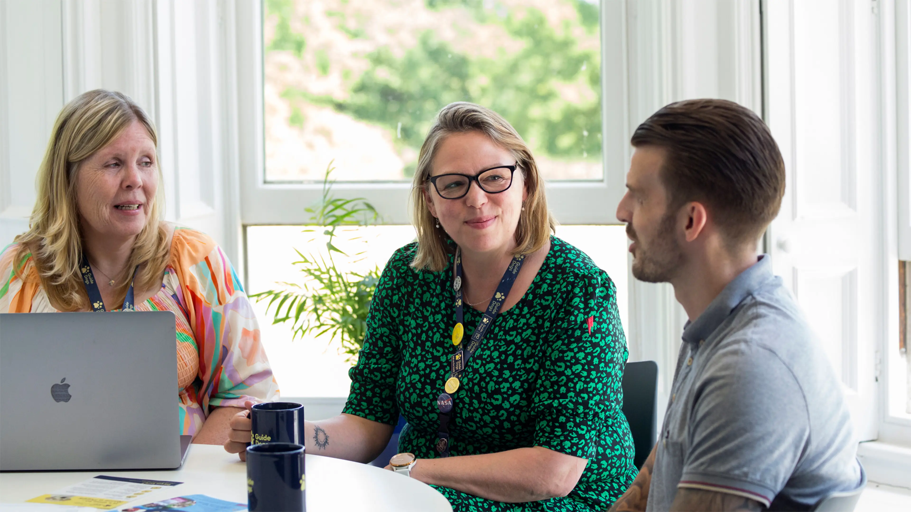 Three Guide Dogs colleagues sit around a table in the office talking over cups of tea.