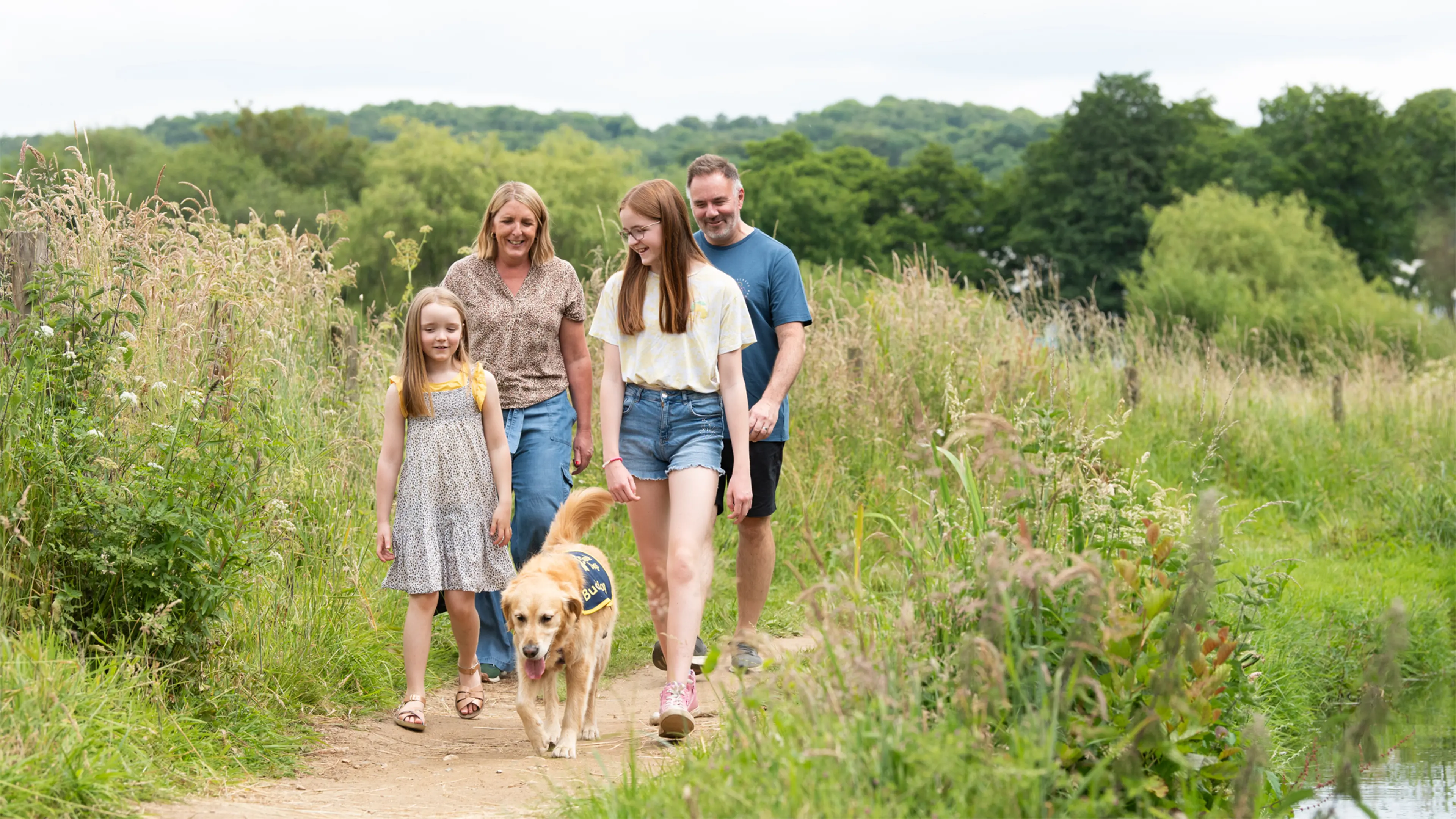Freya, a young person with vision impairment, walks through a field of tall grass with her buddy dog Gwen and her family.